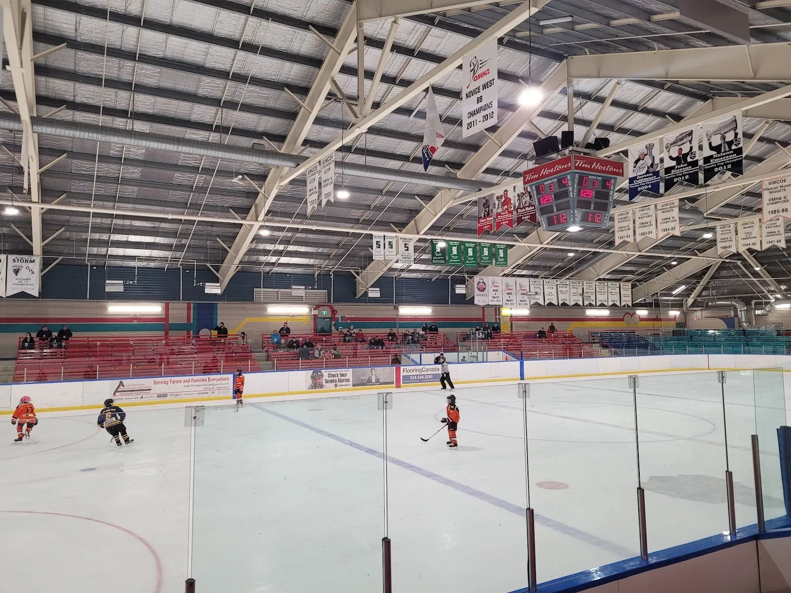 A wide view from the rail across a sheet under a timber-trussed roof, a few players in red on the white ice and banners along the far wall. — Talbot Gardens