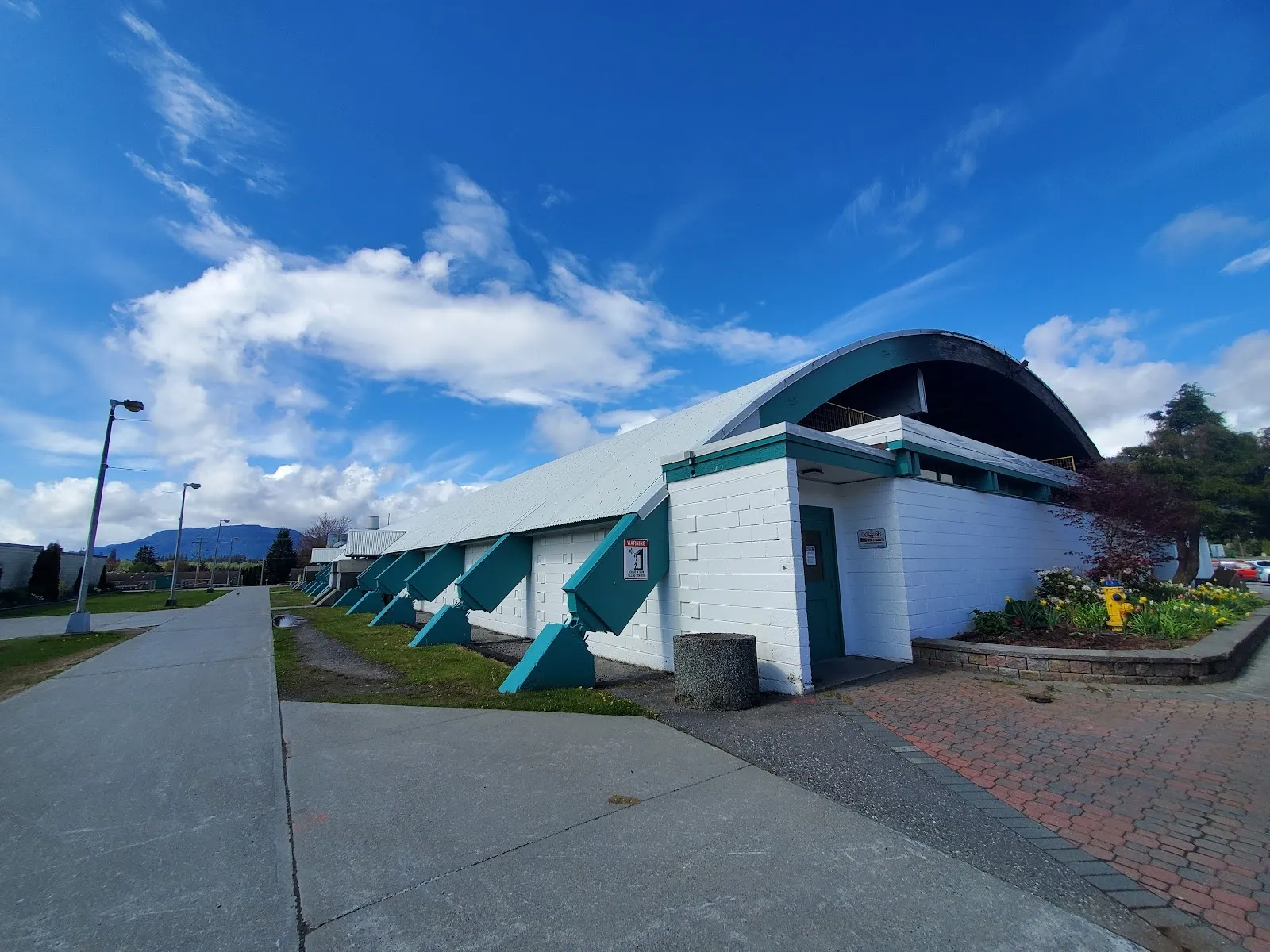 El Tamitik Jubilee Sports Complex, blanco y verde azulado, con un borde de techo curvo y puertas azul verdoso, visto a lo largo de un camino de ladrillo bajo un cielo azul con montañas detrás. — Tamitik Arena