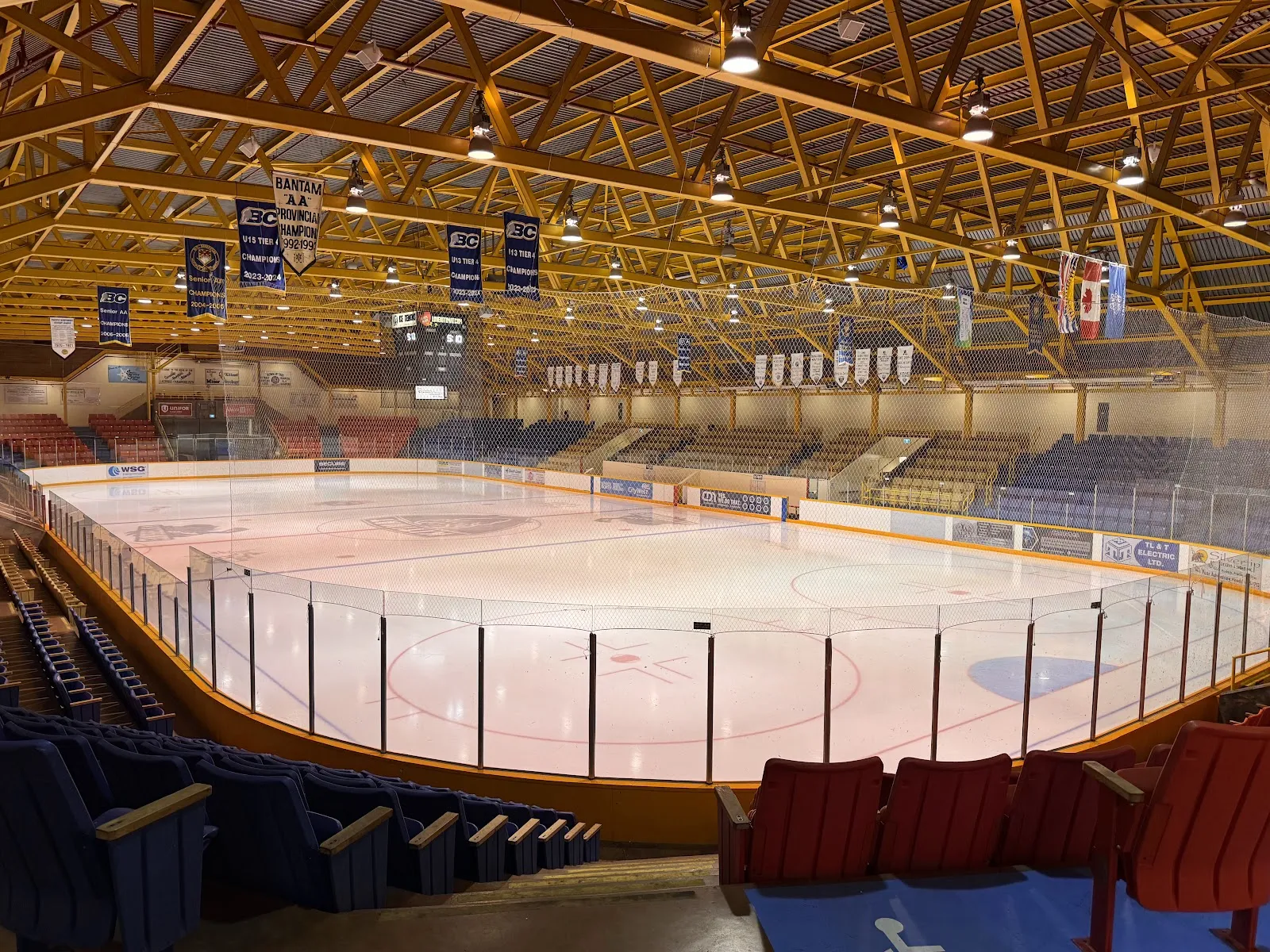 El hielo vacío de Tamitik Arena bajo un entramado de armaduras de madera del que cuelgan mantas de campeonatos, con los asientos elevándose a ambos lados. — Tamitik Arena