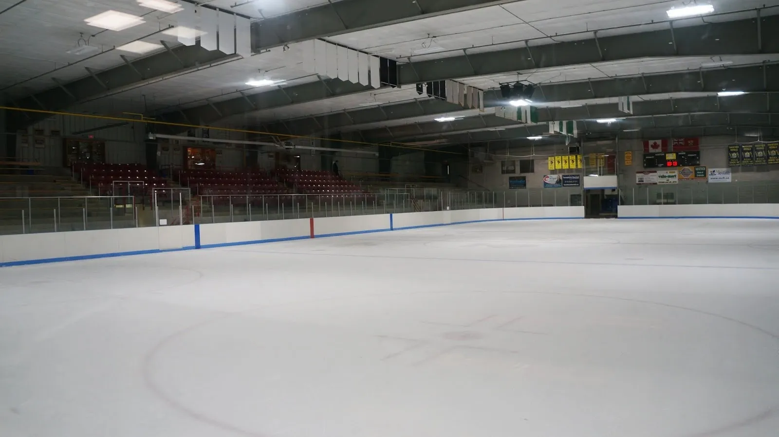 The empty ice sheet at Tavistock and District Recreation Centre from rink level, with the stands, glass and a row of banners along the far side under the arena roof. — Tavistock and District Recreation Centre