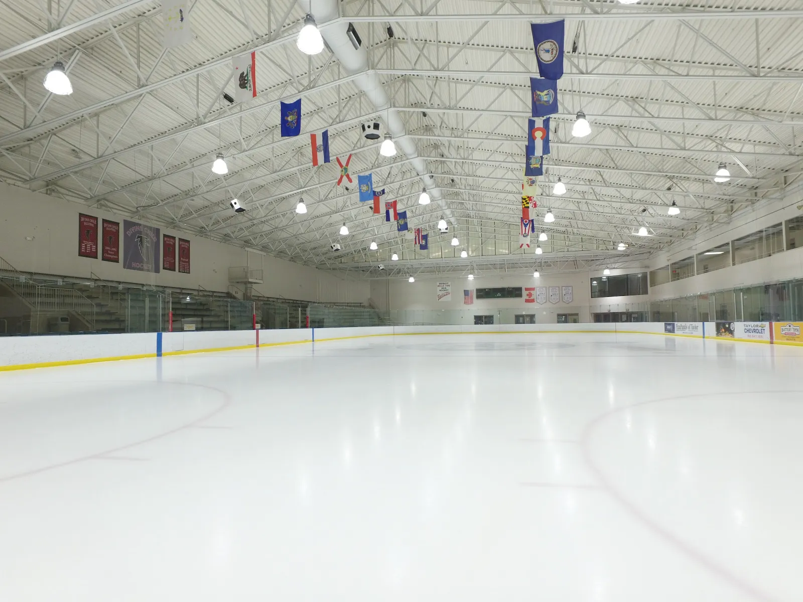 An empty ice sheet with a row of flags hanging from the white steel roof trusses and viewing windows along the far side. — Taylor Sportsplex
