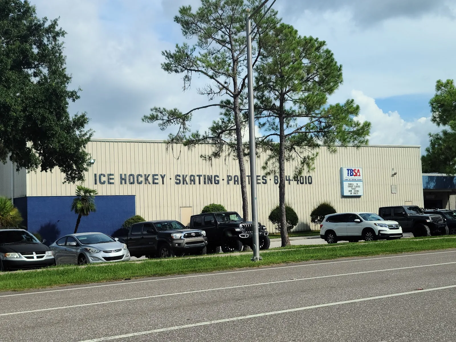 Costado del edificio metálico color beige con el texto pintado "ICE HOCKEY - SKATING - PARTIES - 854-4010" y un letrero de TBSA (Tampa Bay Skating Academy), con autos estacionados a lo largo del muro. — Tampa Bay Skating Academy (Oldsmar)