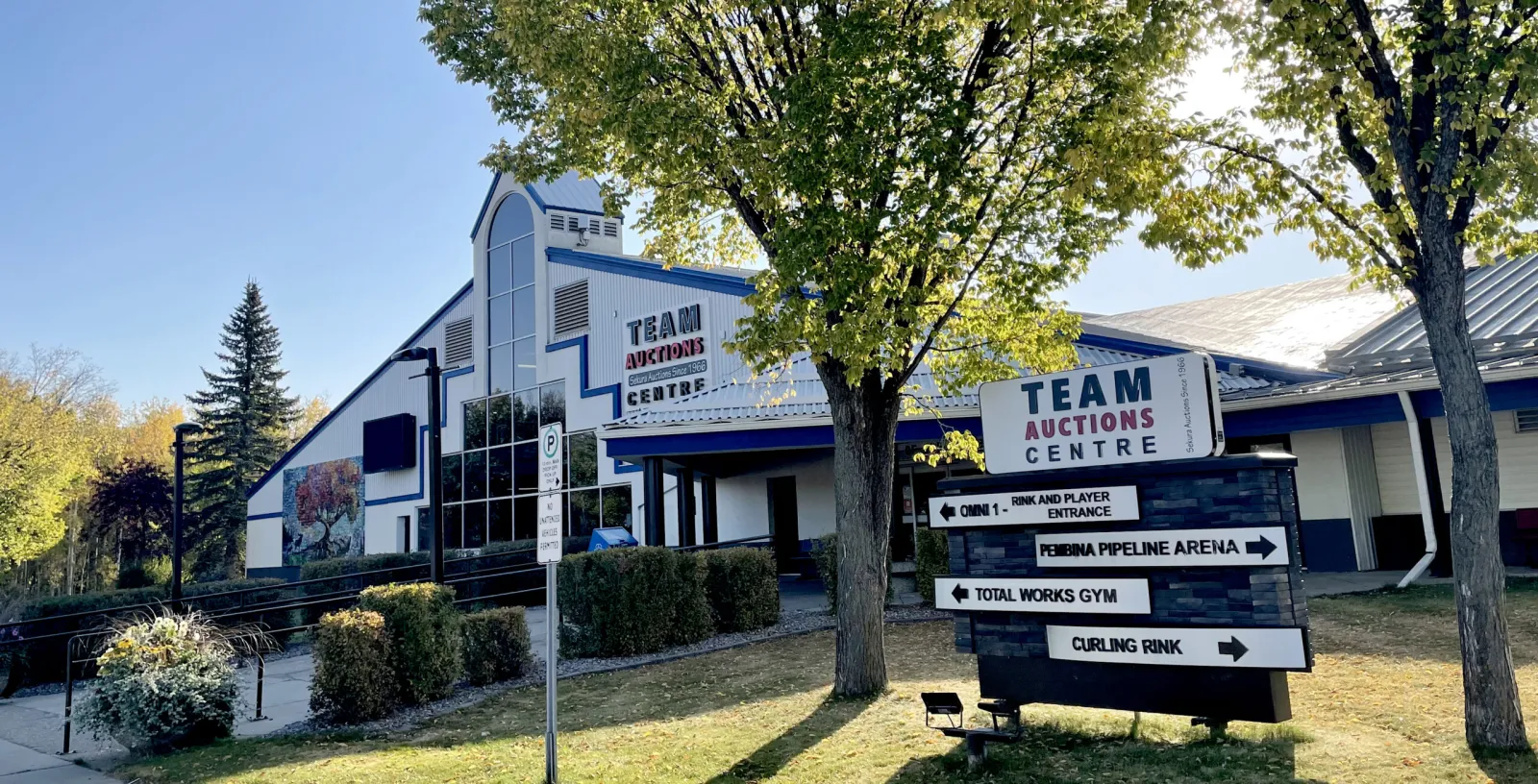 La entrada del Team Auctions Centre, con su nombre en la fachada y un letrero que señala la pista Omni 1, la Pembina Pipeline Arena, el gimnasio y la pista de curling. — Team Auctions Centre