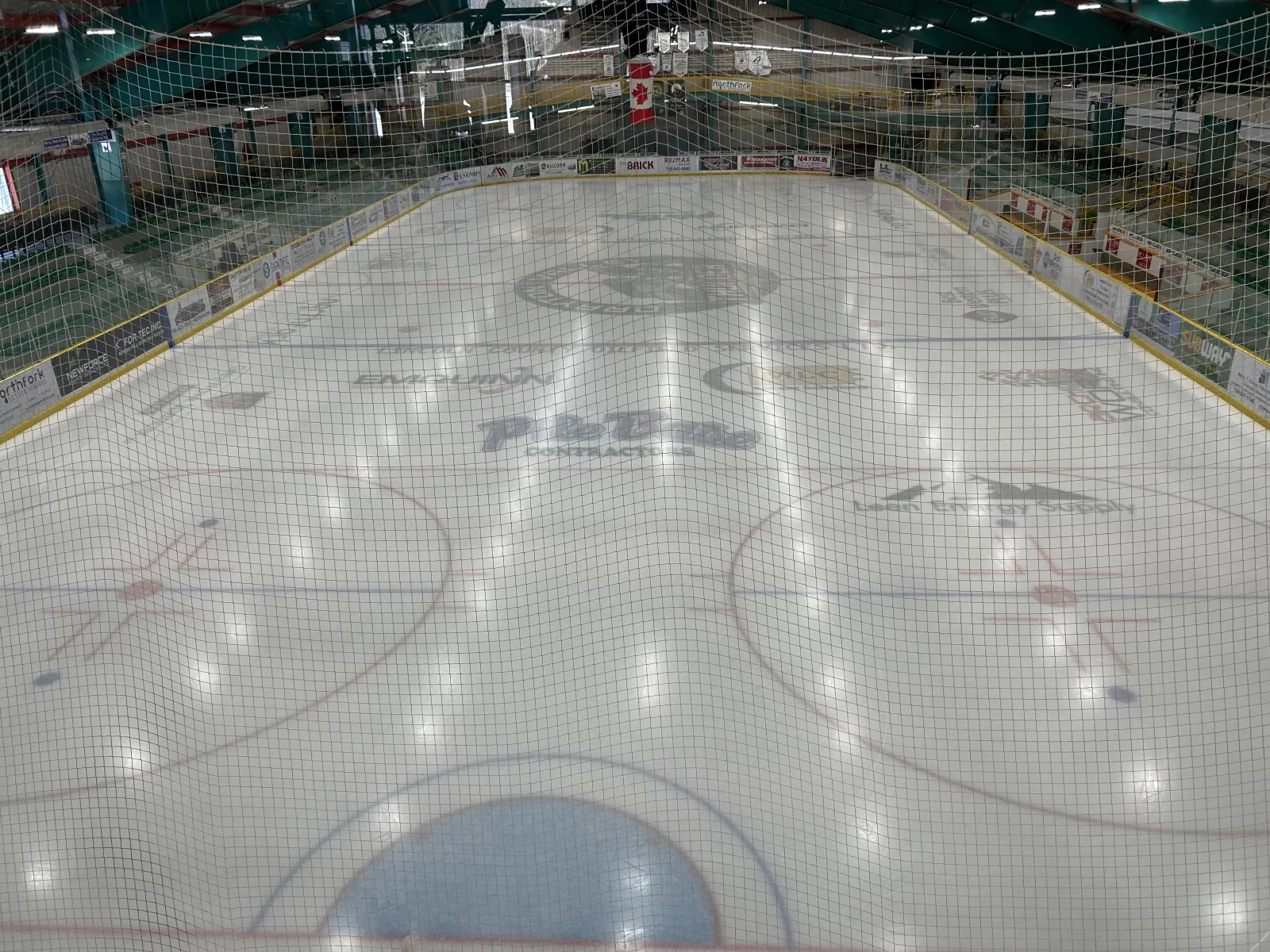 El hielo vacío de la pista Omniplex visto desde detrás de la red de la portería, con logotipos de patrocinadores bajo el hielo y asientos verdes a ambos lados. — Team Auctions Centre
