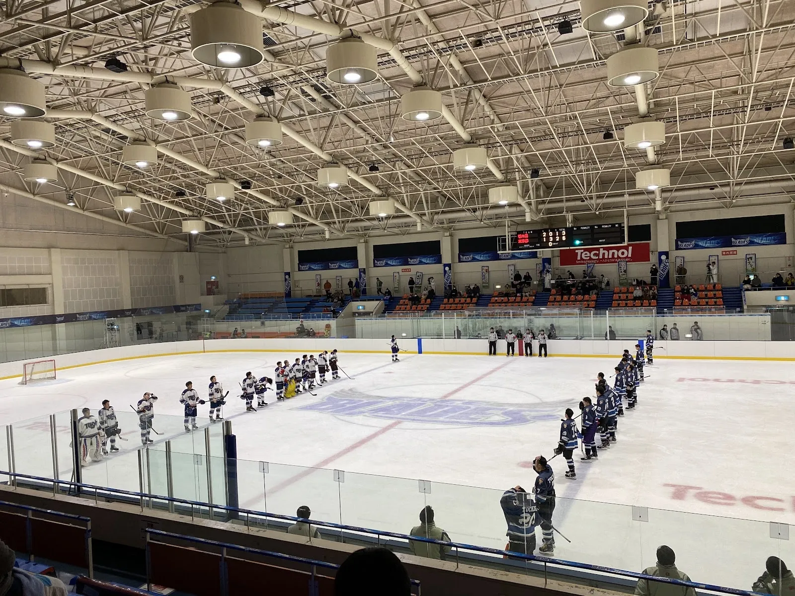 The ice of Technol Ice Park Hachinohe from the stands, with a skating group on the ice and the long arched roof and stands behind. — Technol Ice Park Hachinohe