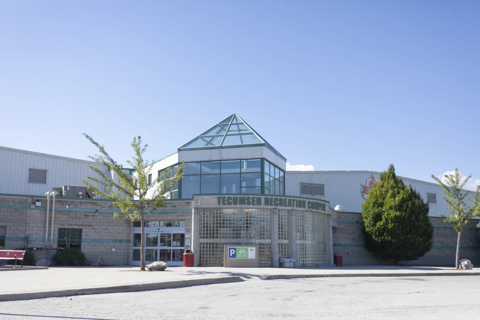La entrada del Tecumseh Recreation Complex: un techo piramidal de vidrio sobre un vestíbulo de bloques de vidrio con el nombre del complejo sobre las puertas, muros de bloques grises y un letrero de estacionamiento al frente. — Tecumseh Arena