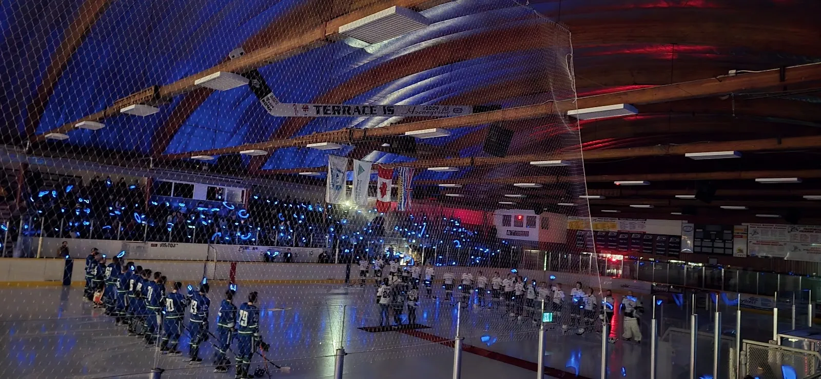 The arena from the stands at an event, a team lined up under blue lighting and timber roof beams above the sheet. — Terrace Sportsplex