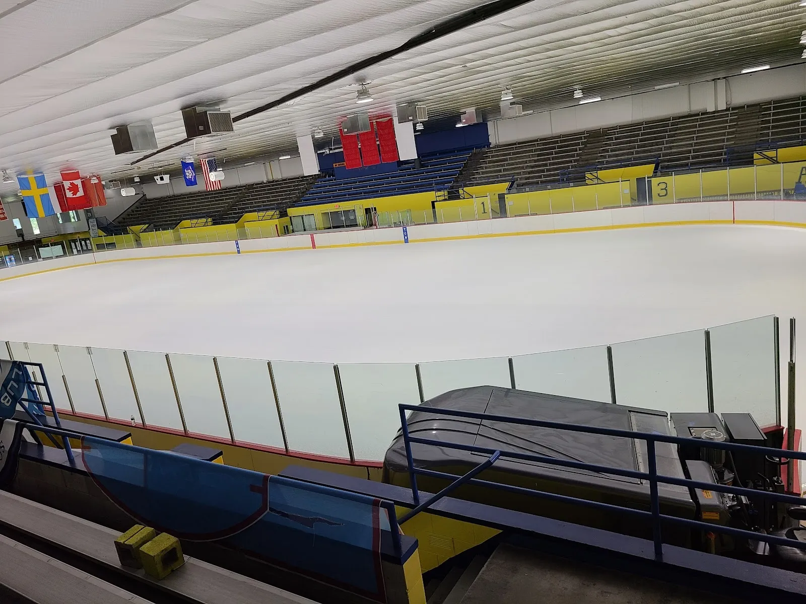 The sheet seen from the glass at the end, banners hung under a sloping ceiling above blue and yellow boards with a bench on the near side. — Terry Conners Rink