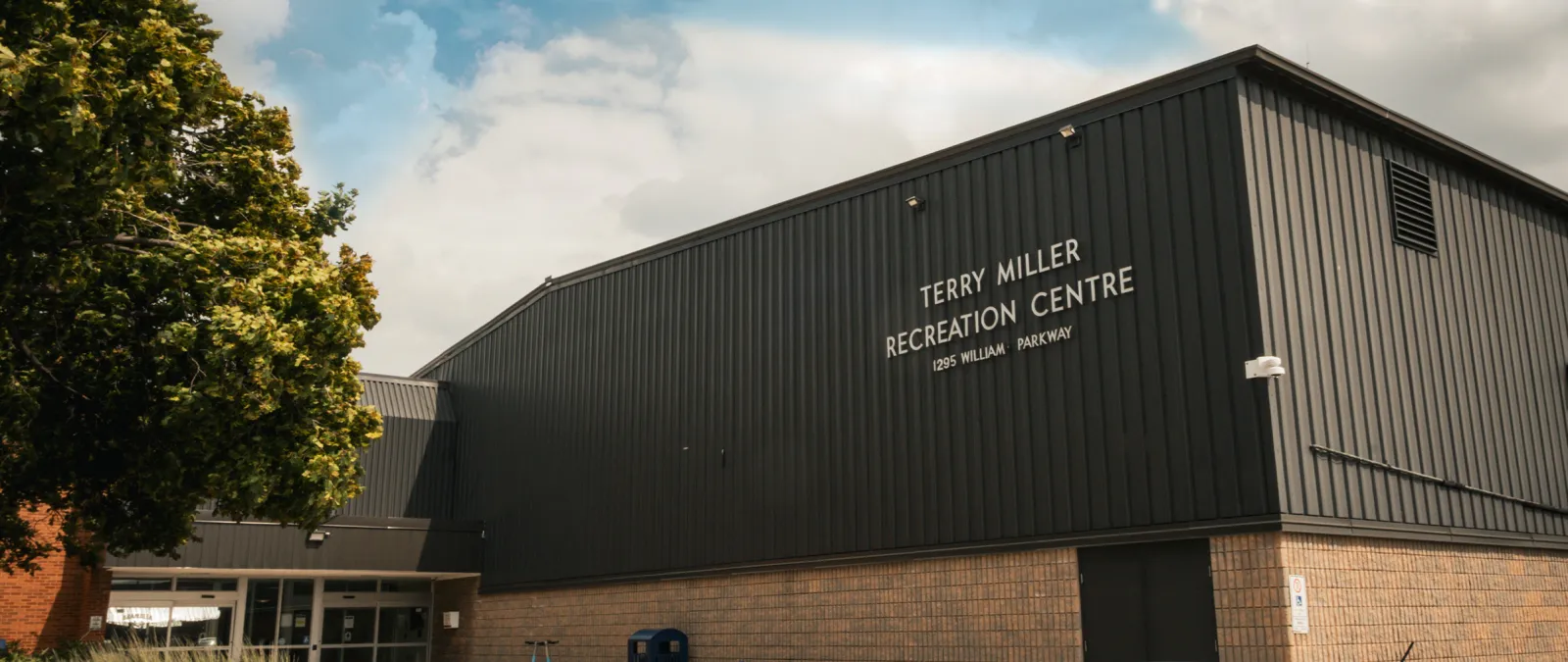 Esquina frontal del edificio de la arena con revestimiento metálico oscuro y base de ladrillo, un árbol a la izquierda y un letrero en la pared que dice "Terry Miller Recreation Centre, 1295 William Parkway" sobre la entrada. — Terry Miller Recreation Centre