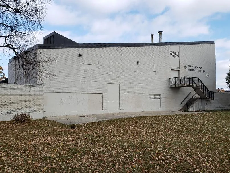 Exterior lateral y trasero de la arena de ladrillo blanco en otoño, con una escalera de emergencia y letreros de "Terry Sawchuk Memorial Arena" en la pared. — Terry Sawchuk Arena