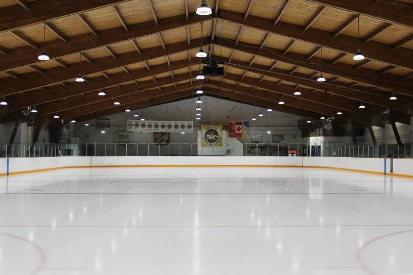 Pista de hielo vacía bajo un techo a dos aguas de vigas de madera, con mantas de equipos y una bandera de Canadá en la pared del fondo. — Terry Sawchuk Arena