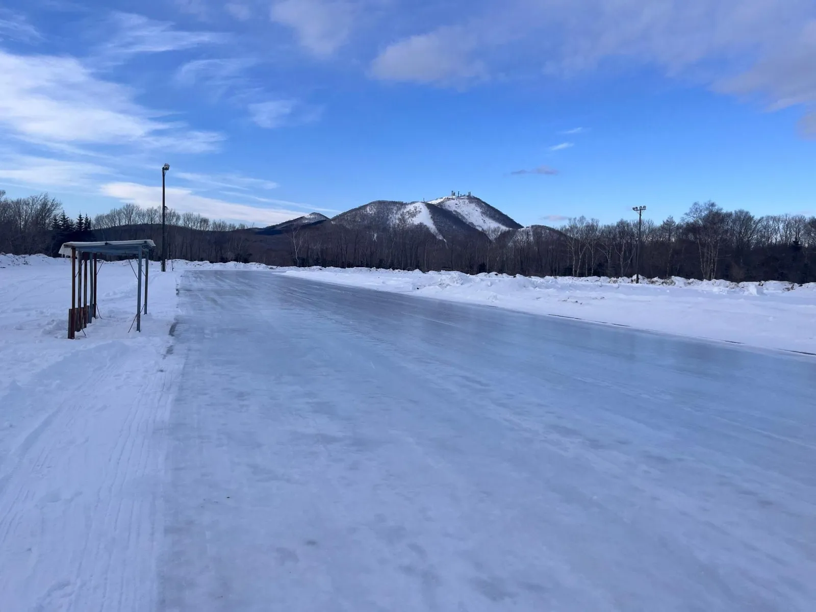 The frozen oval of the Teshikaga town speed skating rink in winter, with the rest shelter and a floodlight beside the track and snowy hills behind. — Teshikaga Town Speed Skating Rink