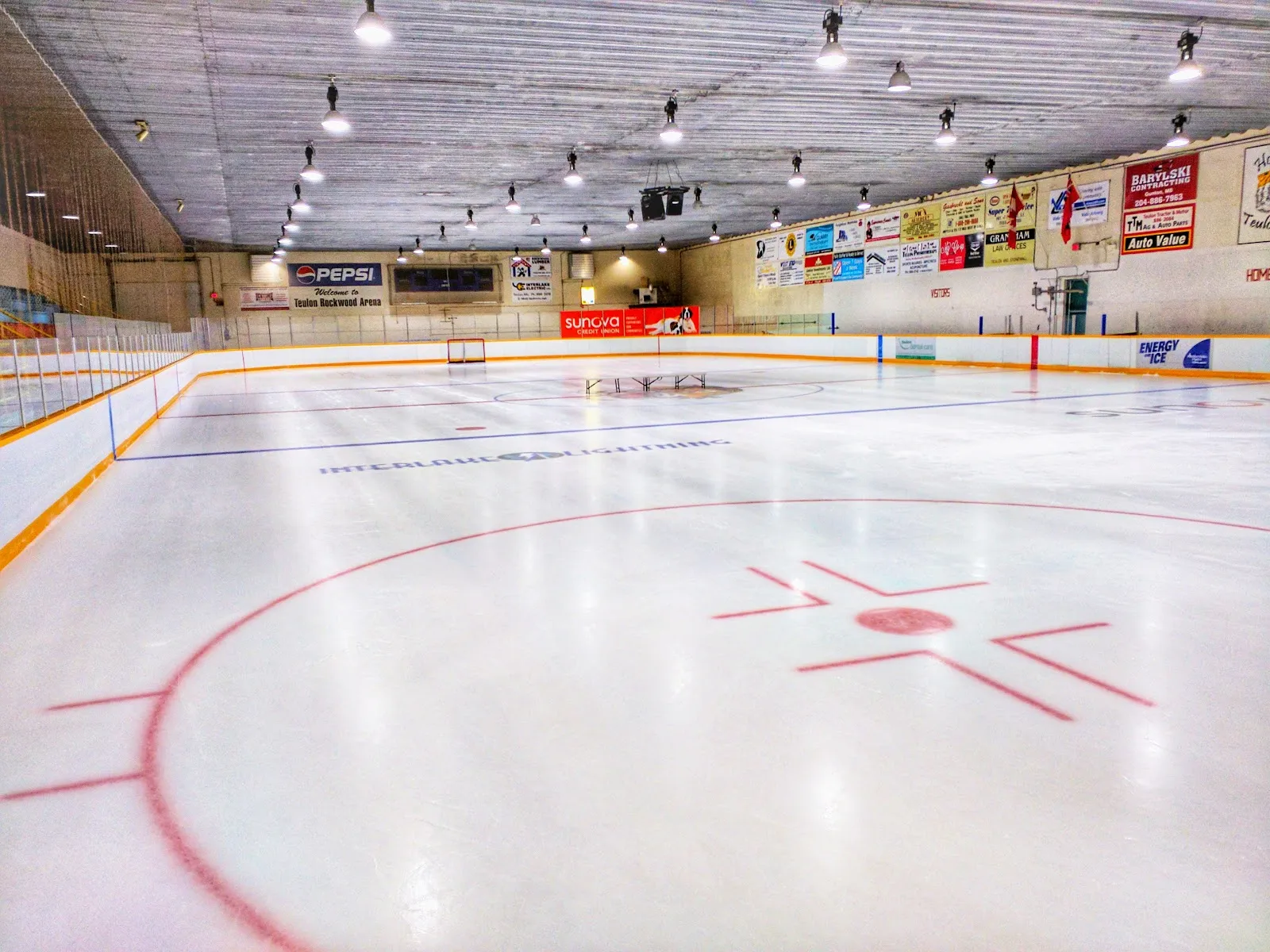 The empty sheet from a faceoff circle, with sponsor boards along the far wall and a wood-panelled ceiling lined with lights. — Teulon-Rockwood Arena