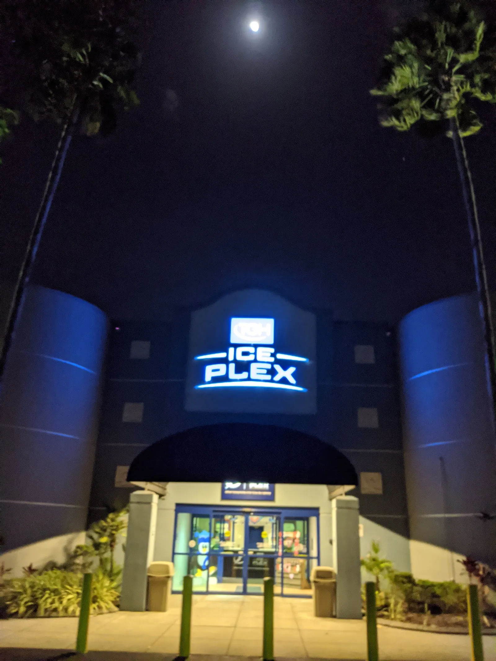 The lit entrance of TGH Ice Plex at night, with the Ice Plex sign above the doors between two round towers and palms either side — TGH Ice Plex