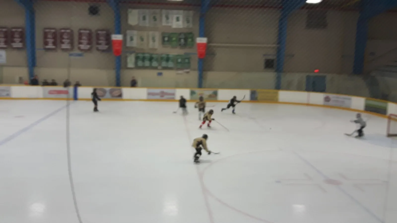 A few players spread across a wide sheet seen through the glass, a grey ceiling and a blue-trimmed wall behind. — Thames Campus Arena