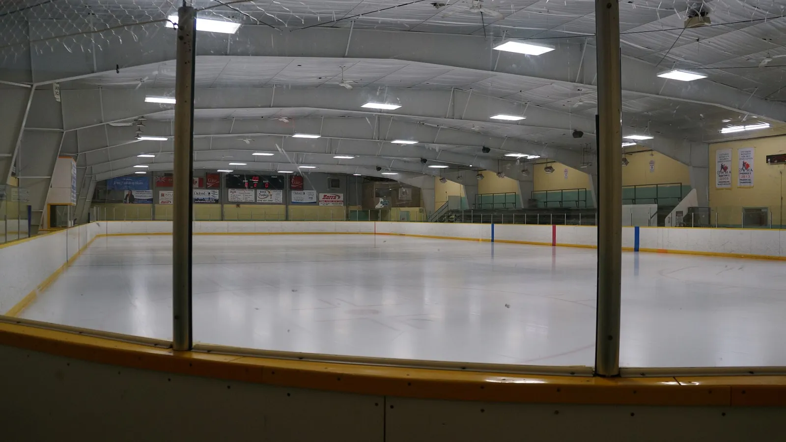 The empty Thamesford arena ice from rinkside, with the yellow-capped boards, low steel roof and sponsor boards along the far end. — Thamesford District Recreation Centre