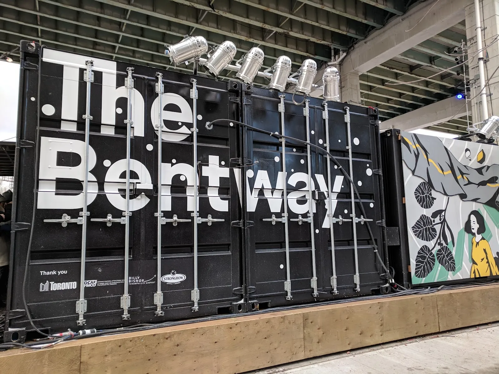 El centro de contenedores marítimos negros de The Bentway con el nombre en grandes letras blancas y un mural al lado, bajo la autopista elevada por donde corre la pista de patinaje. — The Bentway Skate Trail