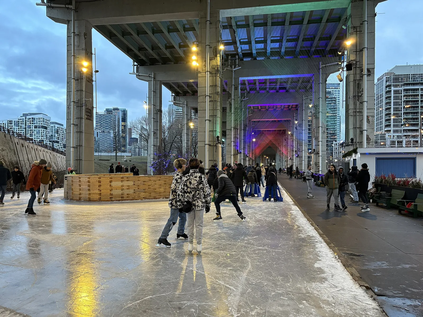 Patinadores en el hielo del sendero de The Bentway bajo la Gardiner Expressway por la noche, con pilares iluminados de colores y torres de condominios de Toronto más allá — The Bentway Skate Trail