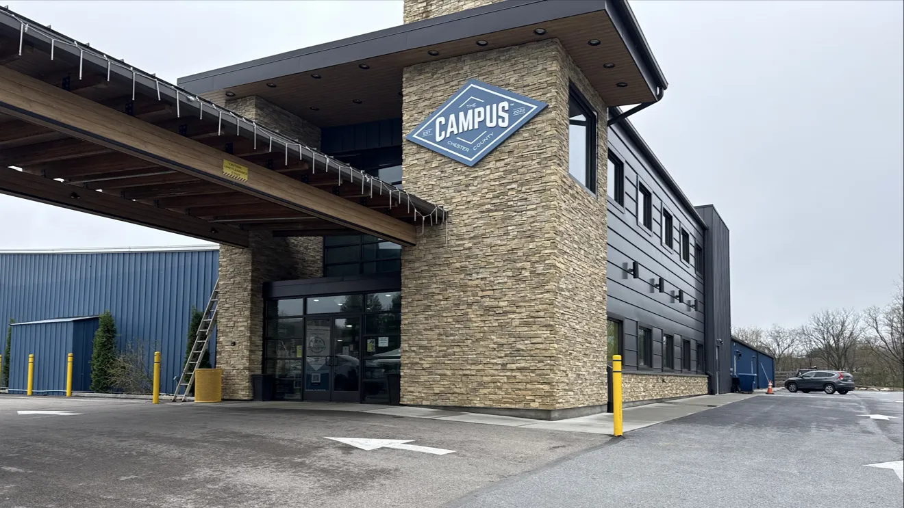 The entrance canopy and stone tower with The Campus sign, beside the long blue metal building and its parking lot. — The Campus