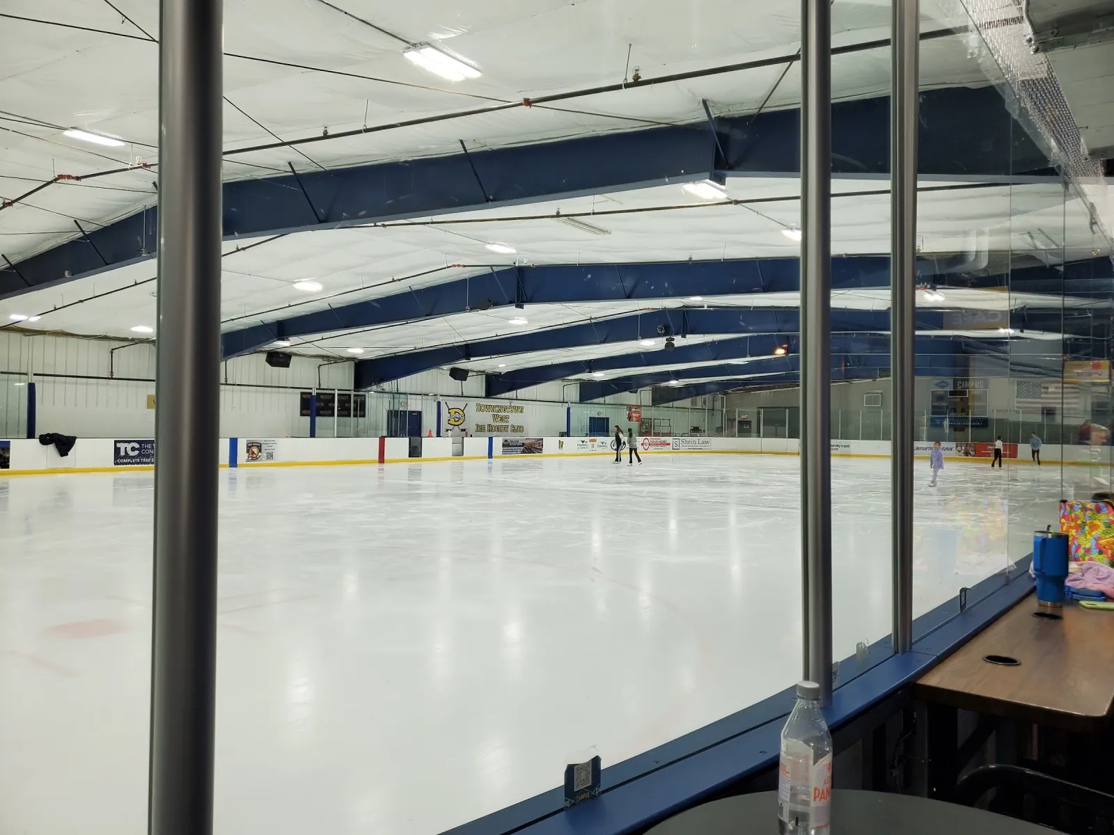 The ice seen through the glass from a table by the boards, with a few skaters at the far end under a white roof with blue beams. — The Campus