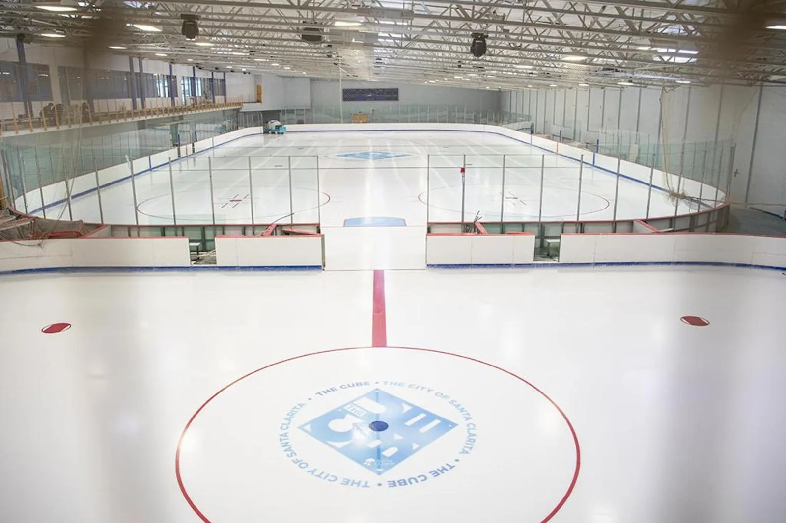 An empty sheet with the Cube logo at centre circle and a red centre line, rink glass and a bright arched ceiling behind it. — The Cube Ice and Entertainment Center