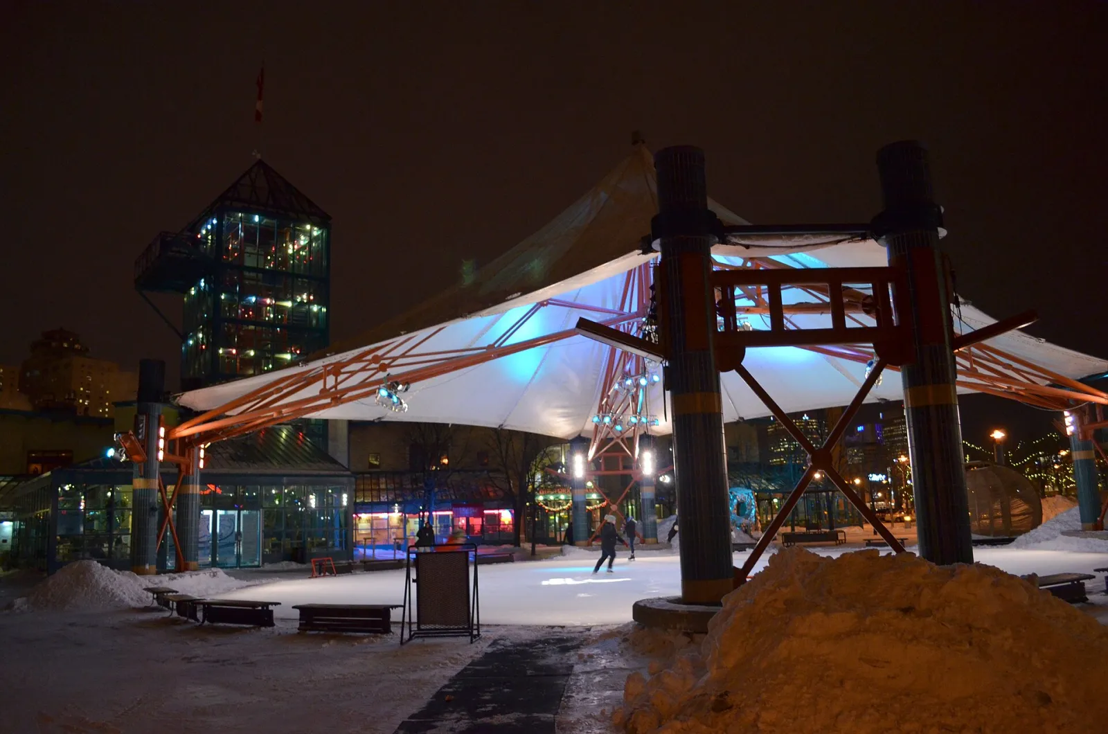 The Forks Winter Park rink under the white canopy at night with a lit tower behind, one distant skater on the ice — The Forks Winter Park Skating Rinks