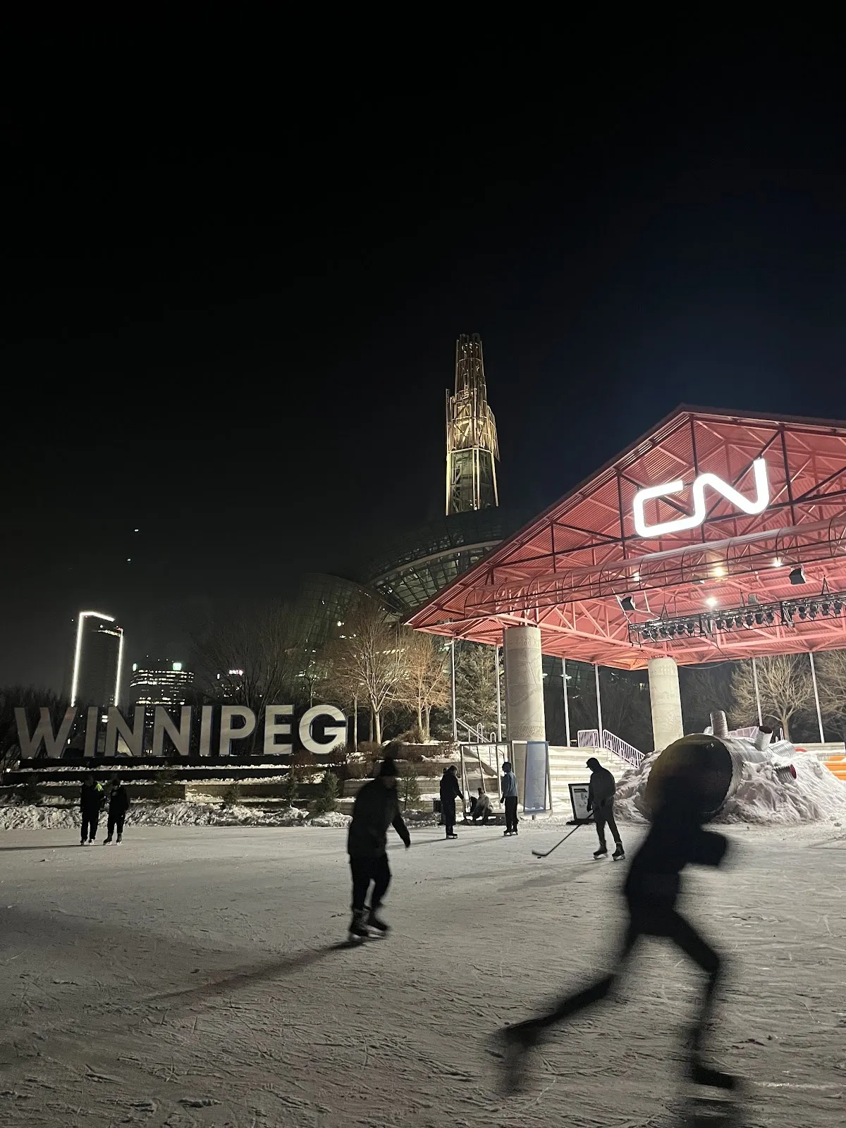 Skaters on the outdoor ice at The Forks at night, with the red-roofed CN Stage pavilion lit behind them and the WINNIPEG letters and a tower in the background. — The Forks Winter Park Skating Rinks