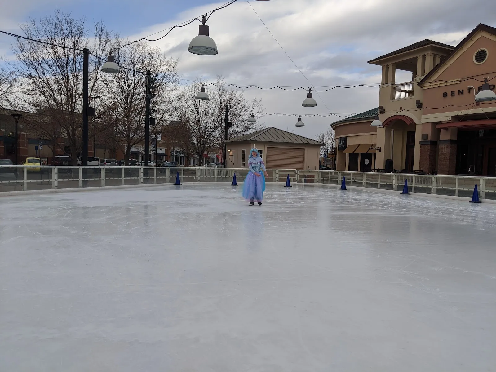 The outdoor rink at The Promenade Shops with a single skater far away and the shopping-centre fronts behind the rail. — The Ice Rink at The Promenade Shops at Centerra