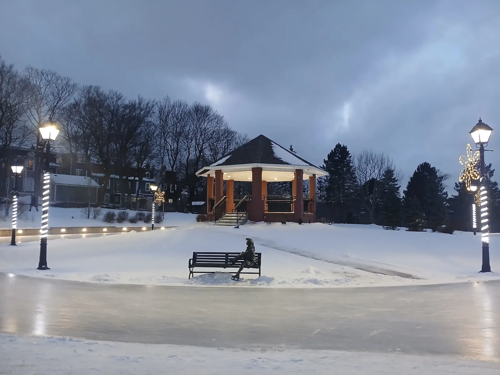 Evening view of the Bannerman Park bandstand lit up beside the frozen, lamp-lit skating loop, with a bench and decorative snowflake lights on the path; the seated figure is a bronze skater statue, not a person — The Loop at Bannerman Park