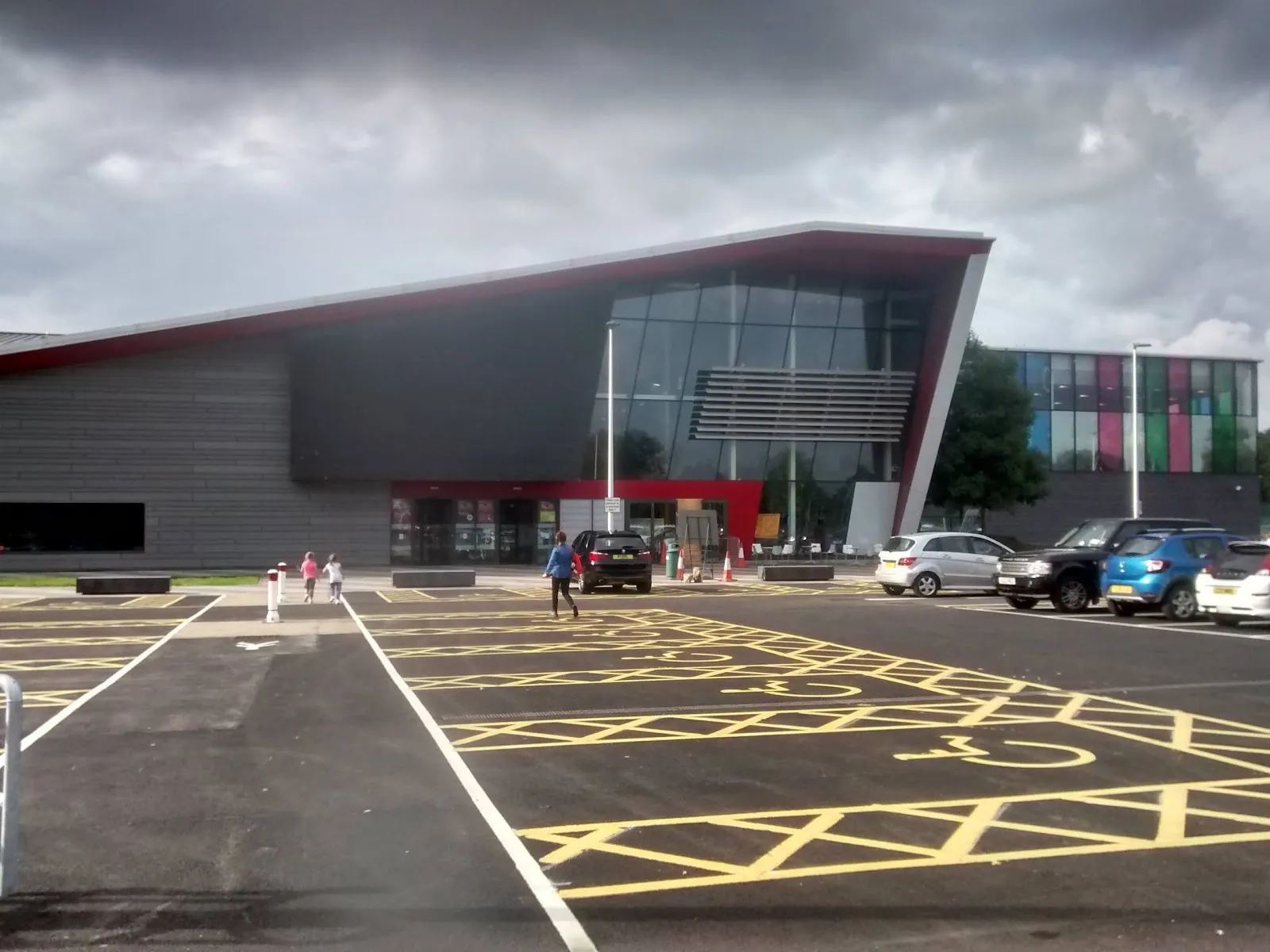 The angular grey and glass front of The Peak sports centre with a red roof edge, seen from the accessible parking bays with hills beyond. — The PEAK Ice Rink