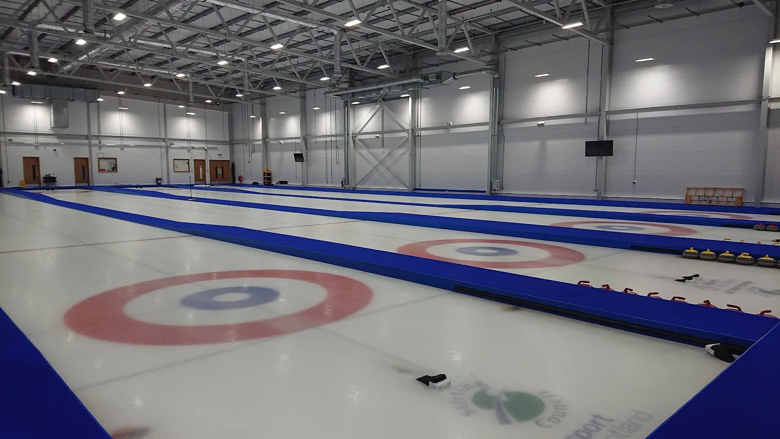 The empty ice hall with curling sheets marked out, a target ring in the foreground and grey cladded walls and steel roof trusses above. — The PEAK Ice Rink