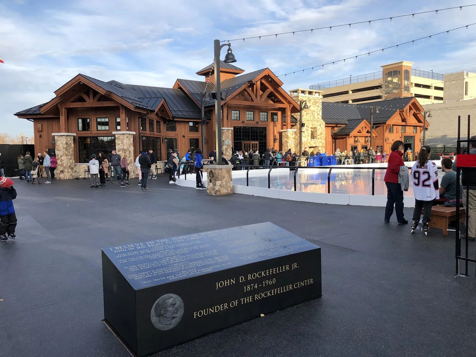 The iced rink in daylight with skaters along the far boards, in front of the timber-and-stone skate house. — The Plaza at Gateway Park