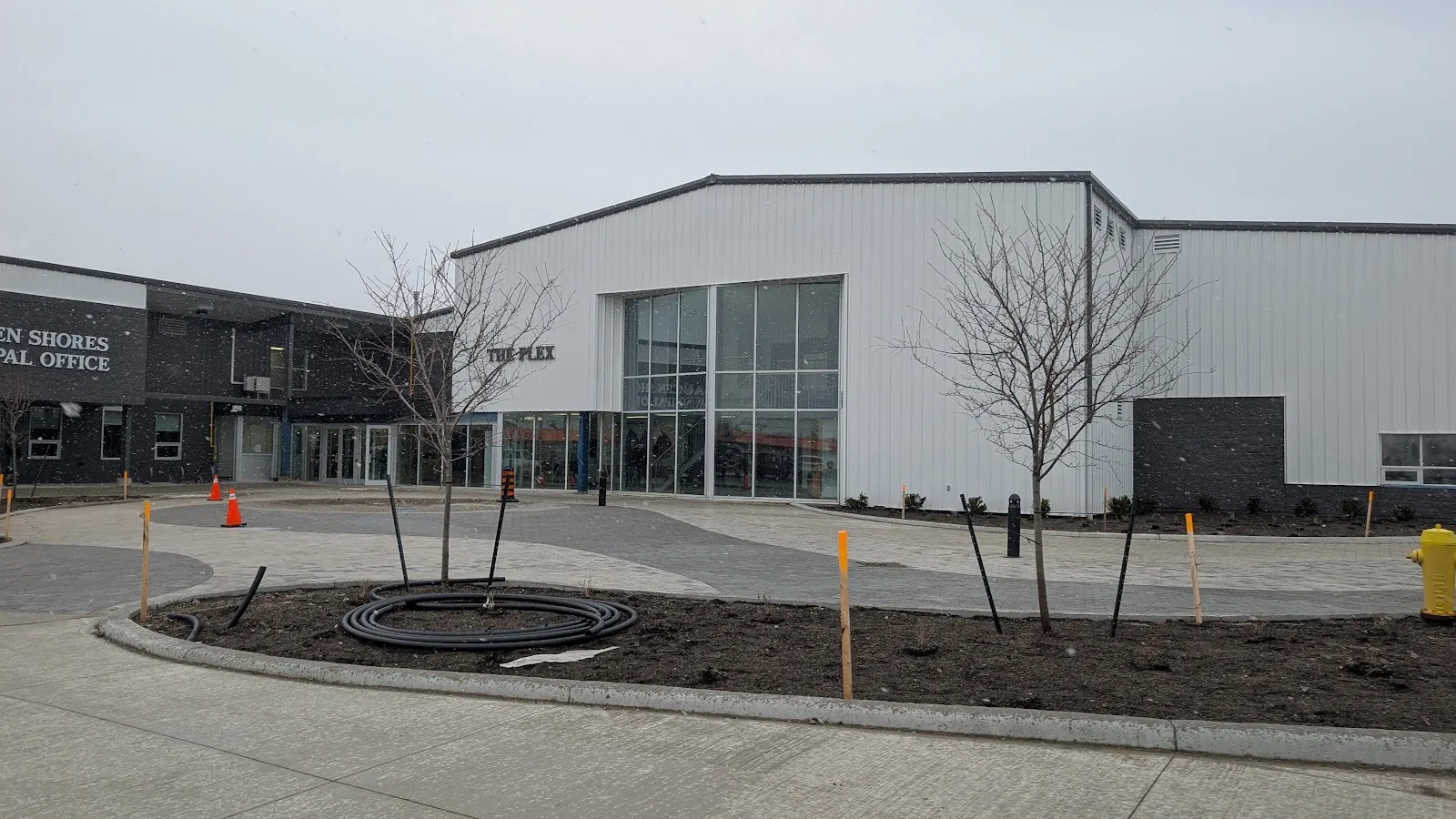 The Plex in Port Elgin, a white metal arena block with tall glazed windows beside the dark-brick Saugeen Shores municipal office, seen from a new paved forecourt with young trees under an overcast sky. — The Plex (Port Elgin)