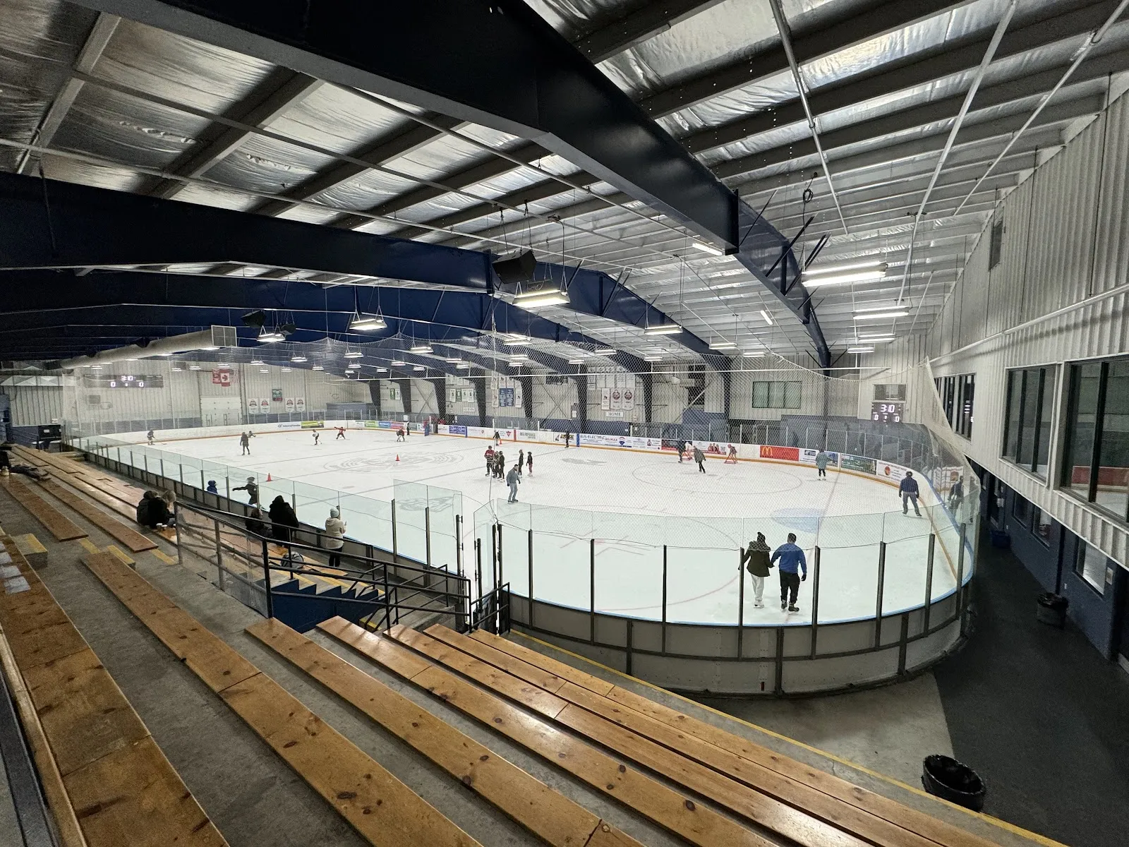 La pista de The Plex vista desde lo alto de las gradas de madera, con un patinaje público en el hielo bajo el techo curvo de acero. — The Plex (Port Elgin)