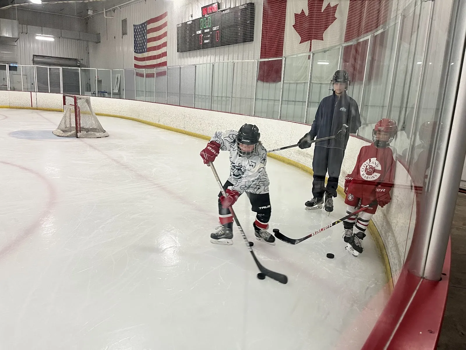 A game seen along the glass, players spread across the near end of the sheet under a wall hung with American and Canadian flags and a score clock. — The Pond Ice Rink & Sports Complex
