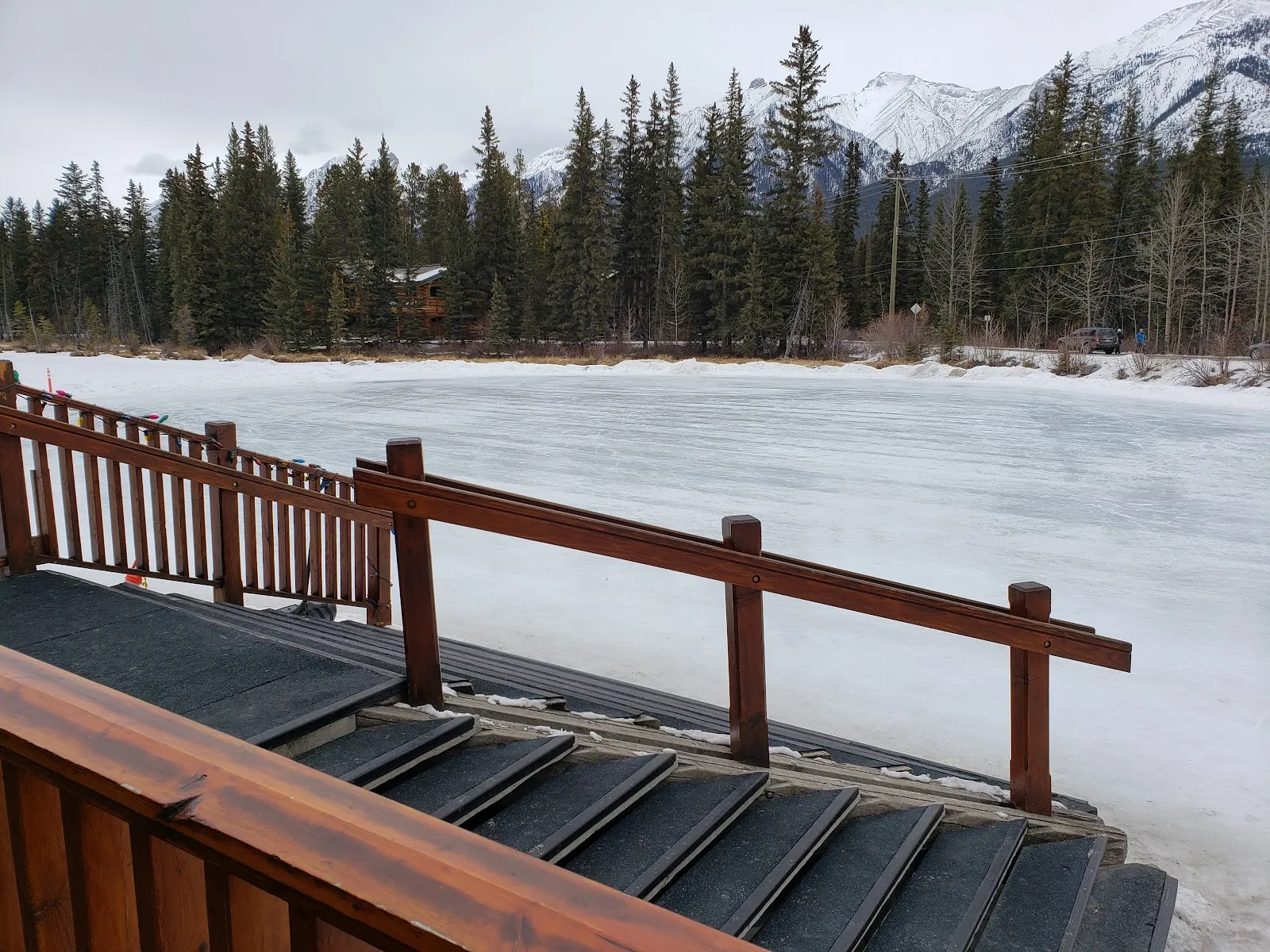 The frozen Canmore pond seen from the wooden boardwalk steps, with spruce trees and the mountains behind. — The Pond Natural Ice Surface (Canmore)