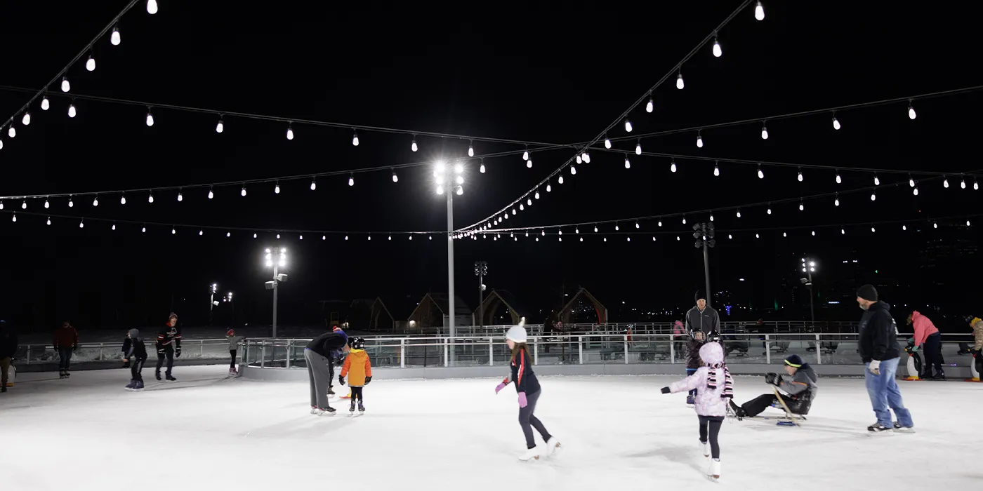 The Pond de noche, bajo tiras de luces, con patinadores y un trineo adaptado en el hielo y las cabañas detrás de la barandilla. — The Ribbon at Glass City Metropark