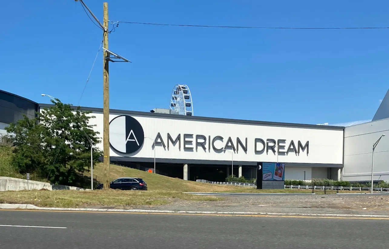 El edificio de American Dream visto desde la Route 3 al otro lado de la carretera, con su gran letrero AMERICAN DREAM sobre el nivel de la entrada y la rueda de observación detrás. — The Rink @ The Arena at American Dream