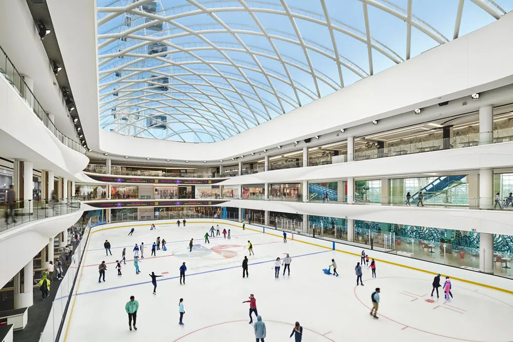 Una sesión pública en la pista vista desde un nivel superior: patinadores repartidos por la pista de tamaño NHL bajo el techo de vidrio del atrio, con los niveles del centro comercial elevándose a los lados. — The Rink @ The Arena at American Dream