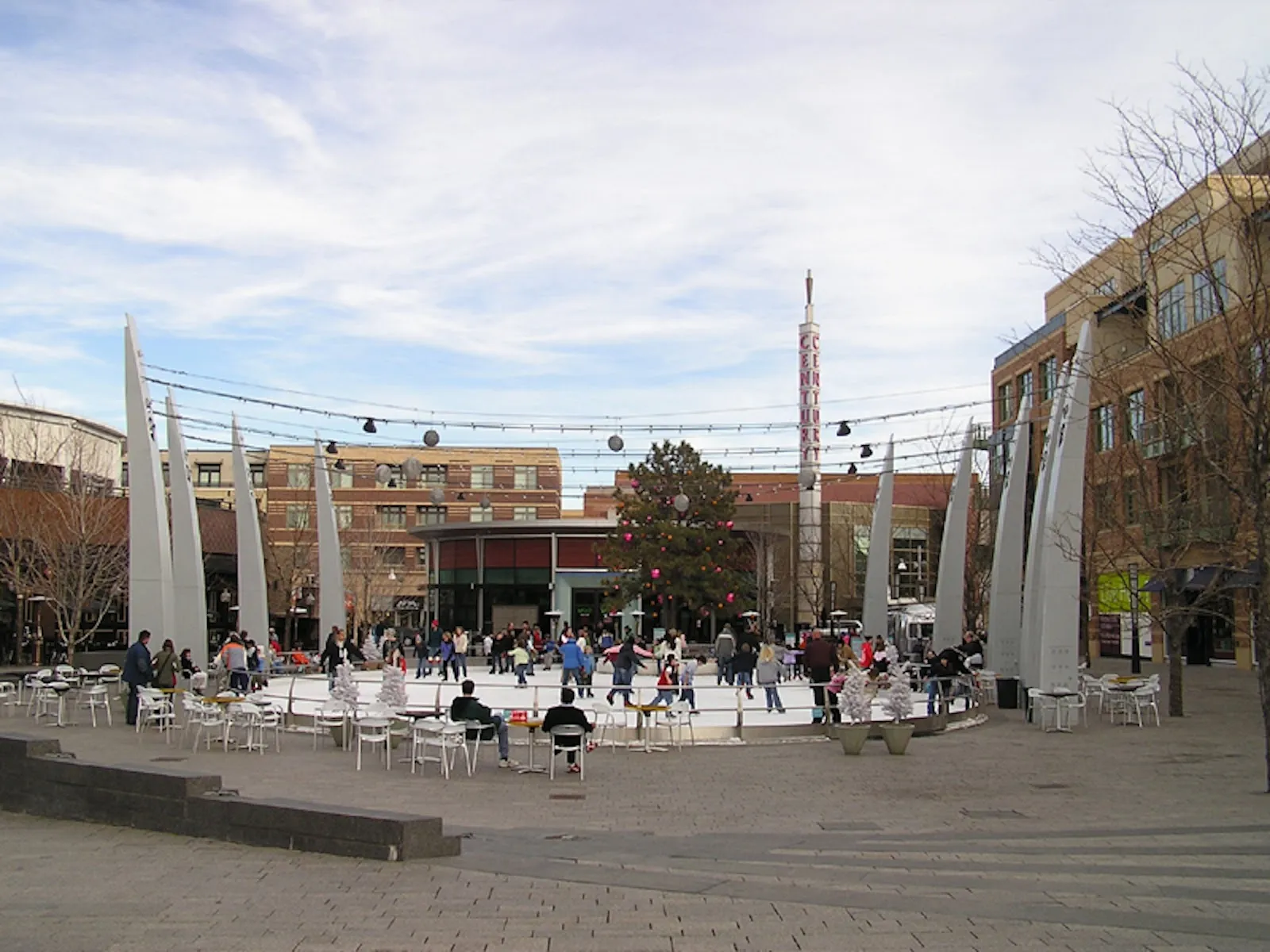 Distant skaters on the plaza rink with the tall light masts and storefronts around it — The Rink at Belmar