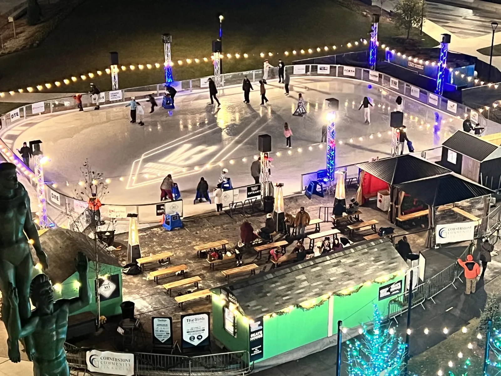 La pista de noche desde arriba, rodeada de tiras de luces, con la casa de patines y las cabañas para entrar en calor a un lado y patinadores dando vueltas por el hielo. — The Rink at Royal Oak