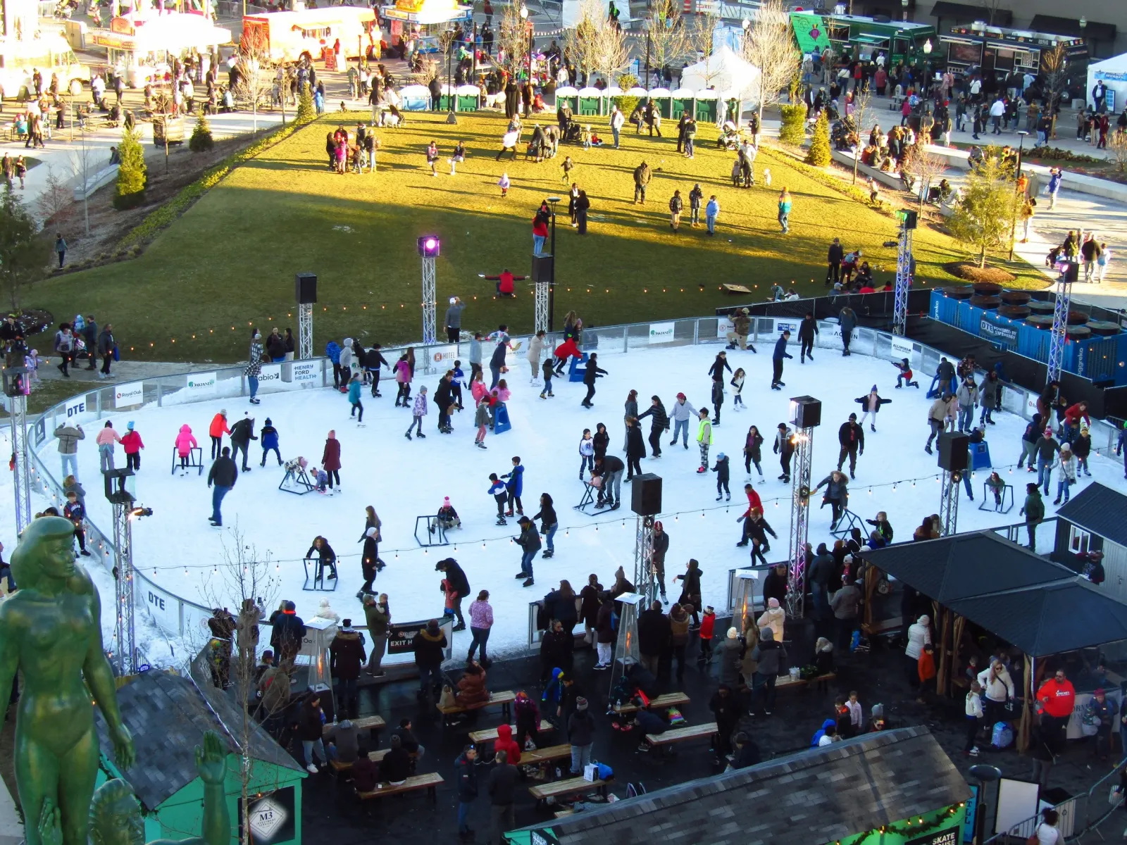 Toda la pista de 60 por 90 pies desde arriba en una tarde concurrida, con el césped de Centennial Commons detrás y la casa de patines en el borde del hielo. — The Rink at Royal Oak
