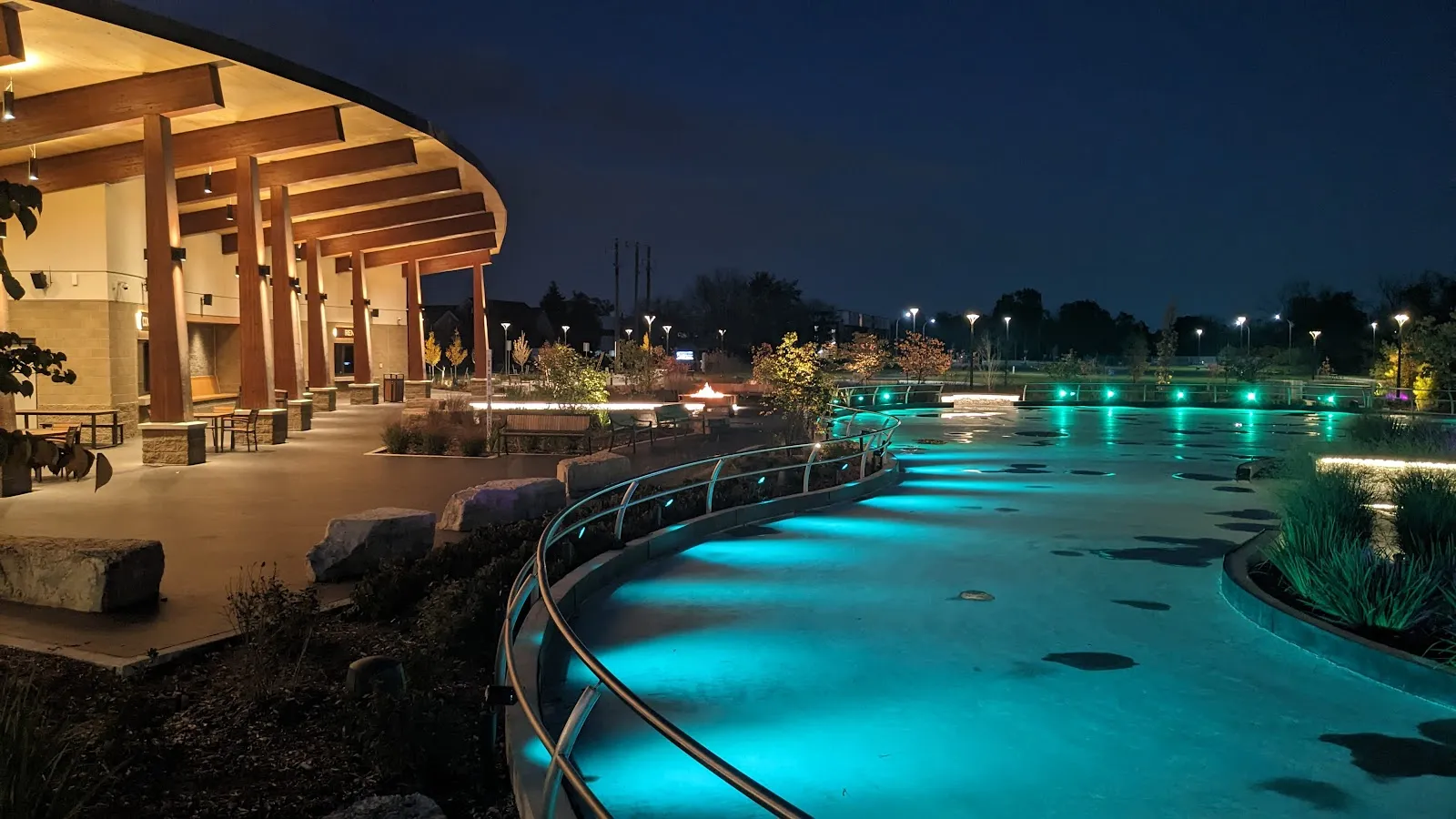 El pabellón de madera con su techo curvo junto a la cinta de patinaje iluminada de noche. — The Rink at Stine Community Park