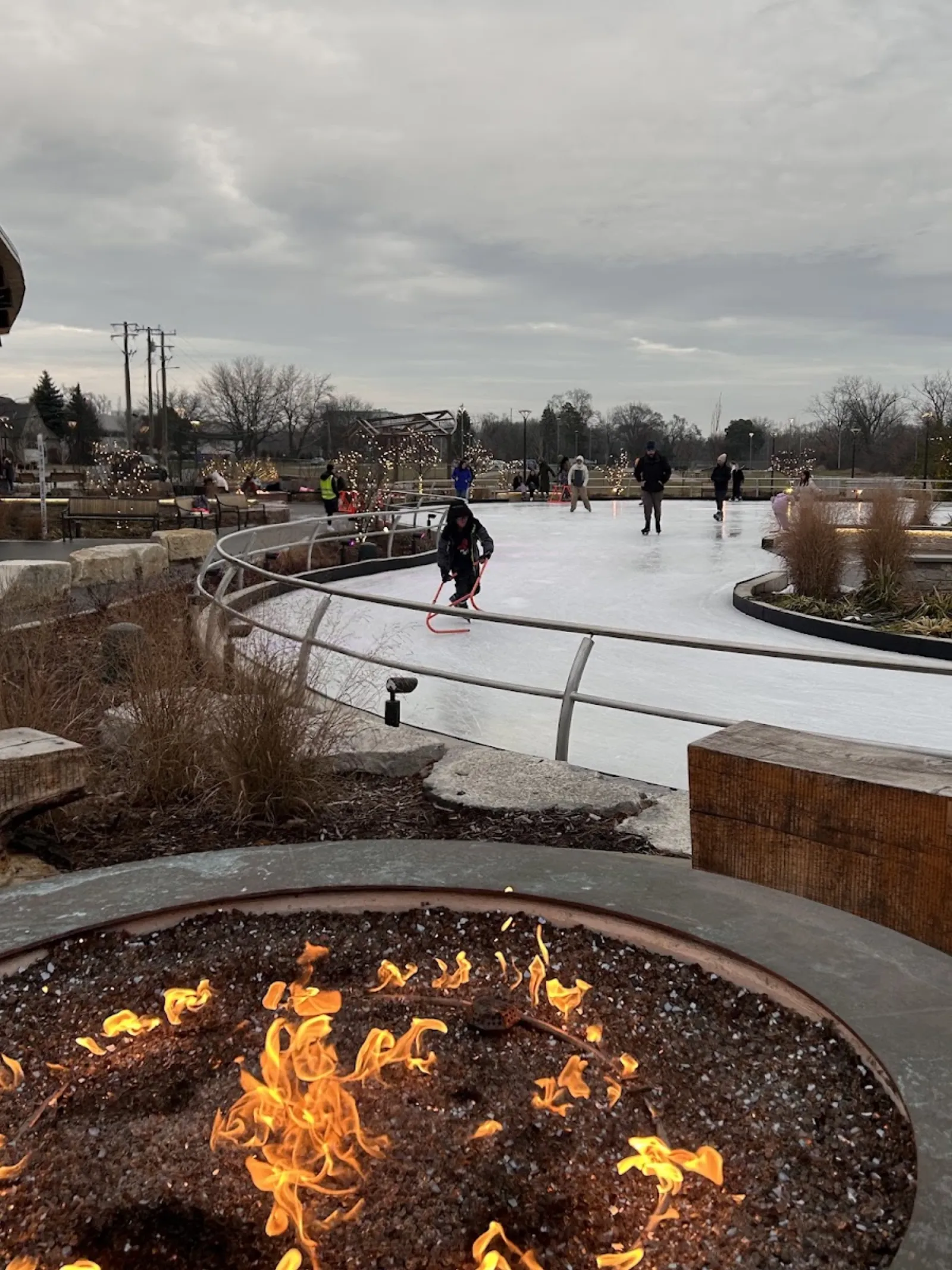 La cinta de hielo curva con su baranda de acero, una fogata en primer plano y algunos patinadores en el hielo. — The Rink at Stine Community Park
