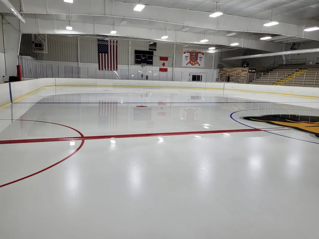 The empty sheet from the corner, with red and blue lines, the flags and bleachers at the far end and bright overhead lights. — The Rink Battle Creek
