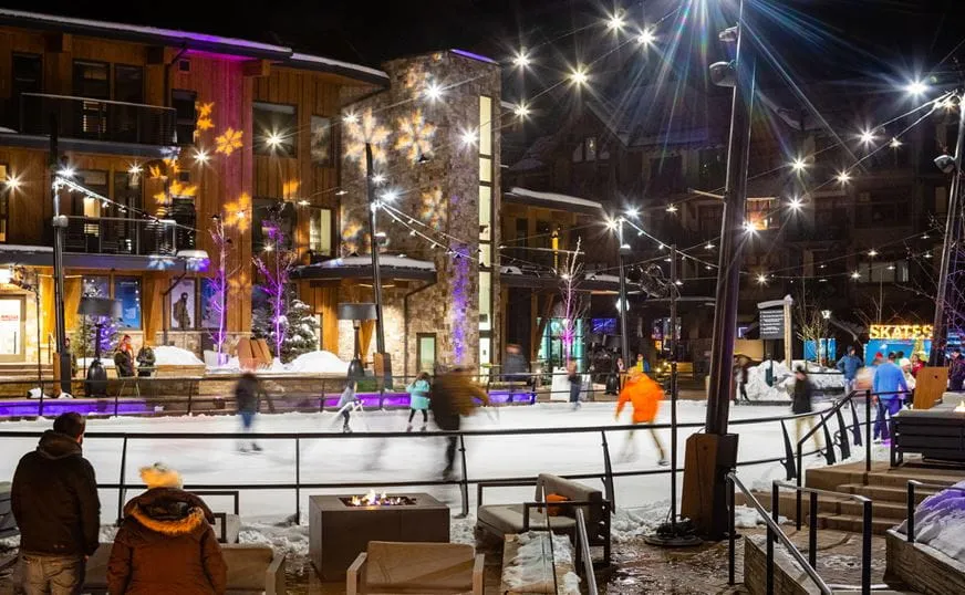 The Rink at Snowmass Base Village in the evening, with the lit ice in front of the lodge buildings and strings of lights overhead. — The Rink at Snowmass Base Village