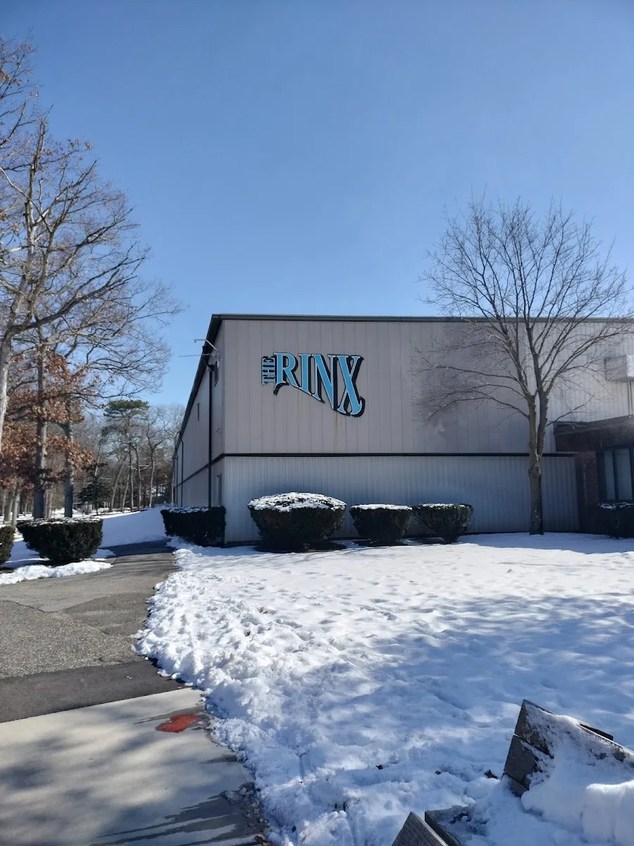 The grey rink building with The Rinx logo high on its wall, seen across a snowy lawn and trimmed shrubs on a clear winter day. — The Rinx