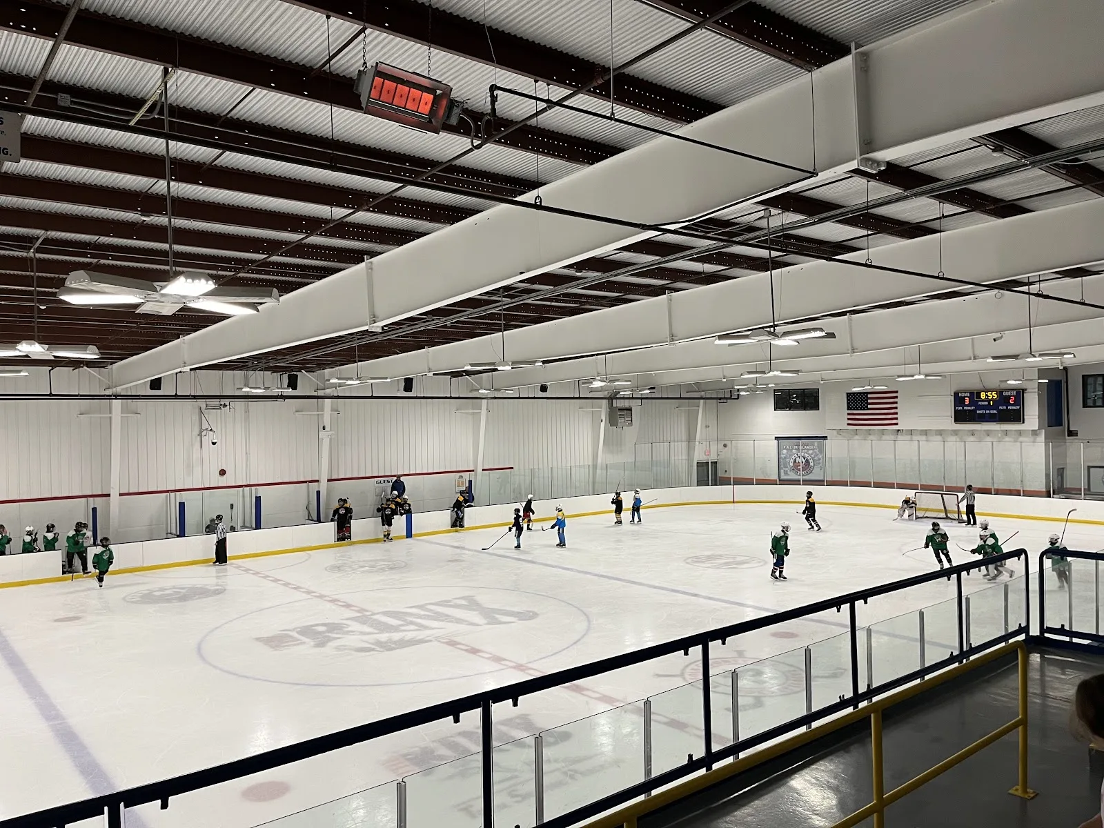 Young skaters in green spread across one of the rinks, seen from the raised walkway above the boards under a white steel roof. — The Rinx