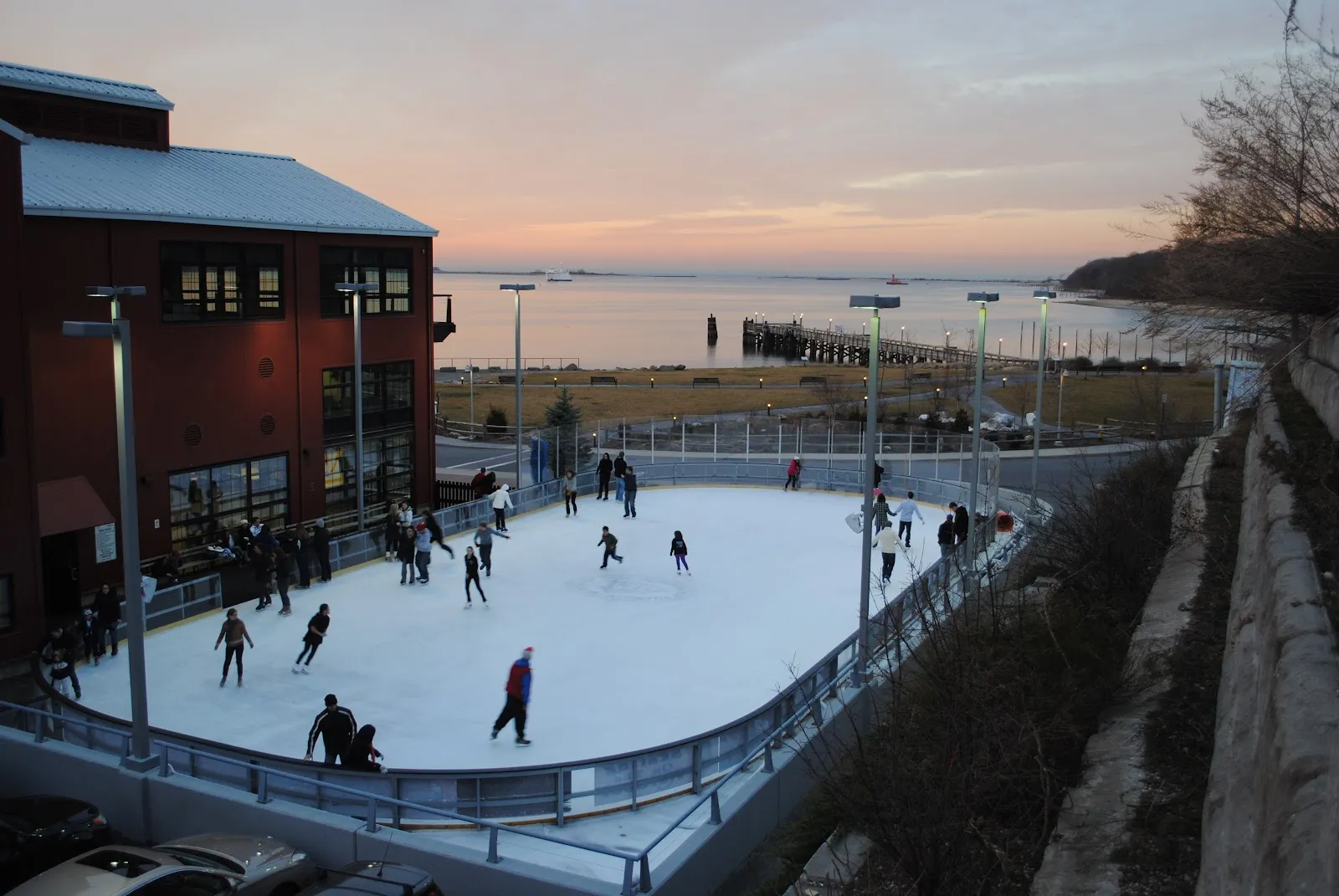La pista exterior al anochecer junto al Port Jefferson Village Center de ladrillo, con patinadores en el hielo y el puerto y un ferri al fondo. — The Rinx at Harborfront Park