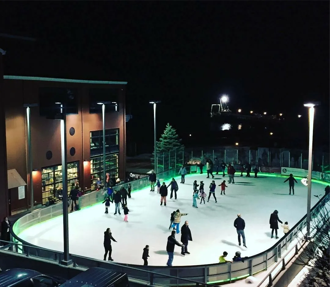 Toda la pista ovalada de noche durante una sesión pública, con un resplandor verde a lo largo de las bandas y el Village Center detrás. — The Rinx at Harborfront Park